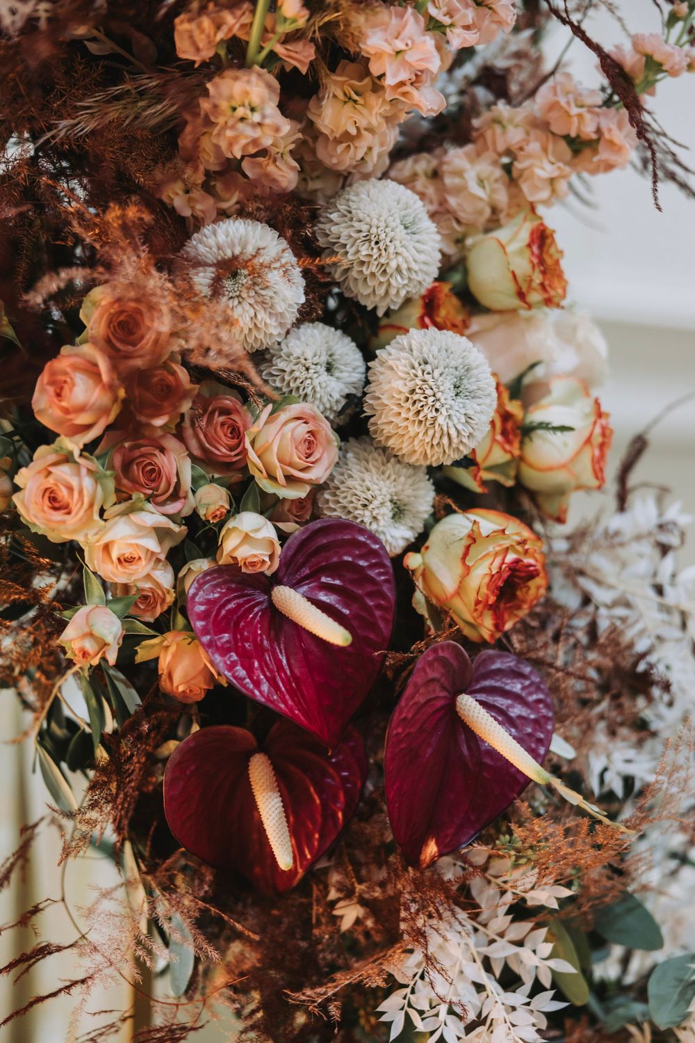 Moody floral arrangement featuring peach roses, white chrysanthemums and deep burgundy anthurium blooms.