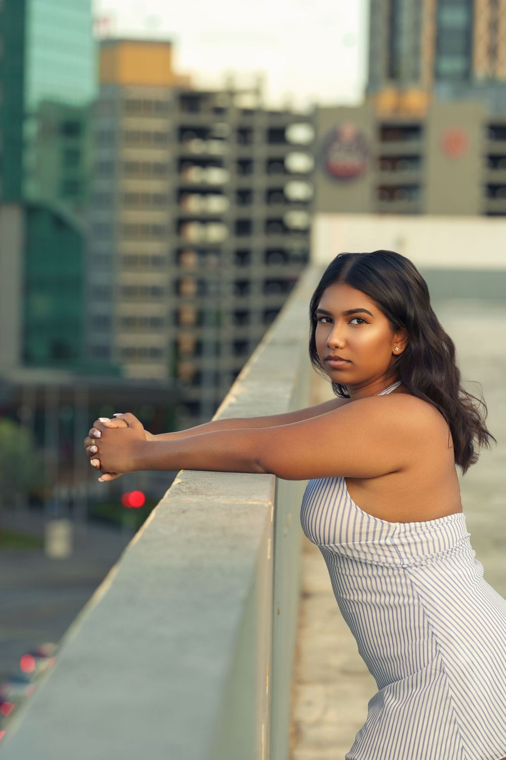 Person in a white striped dress leans against a rooftop railing with city buildings in the background at sunset.