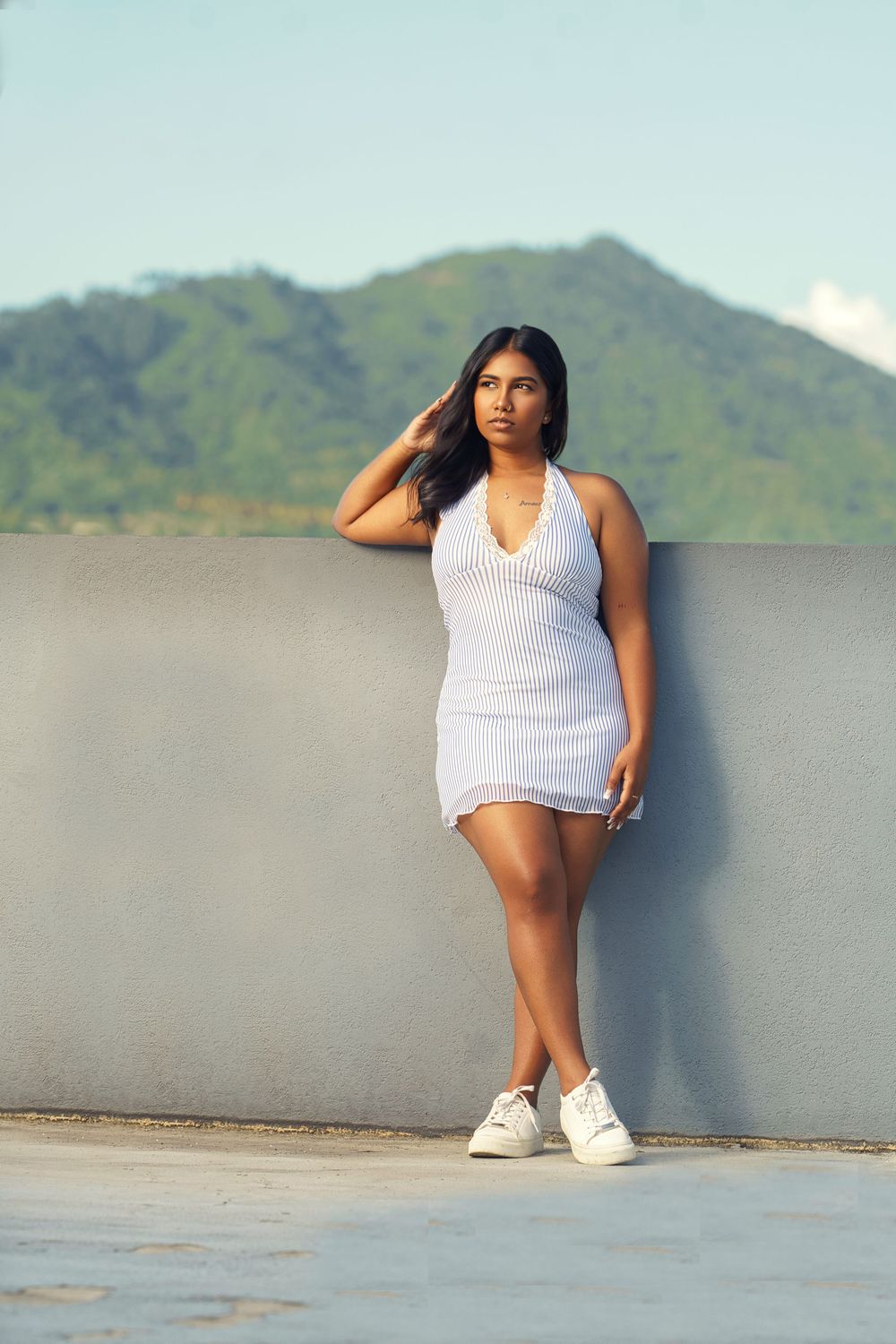 A person in a white tank top poses casually against a gray wall with mountains visible in the background.