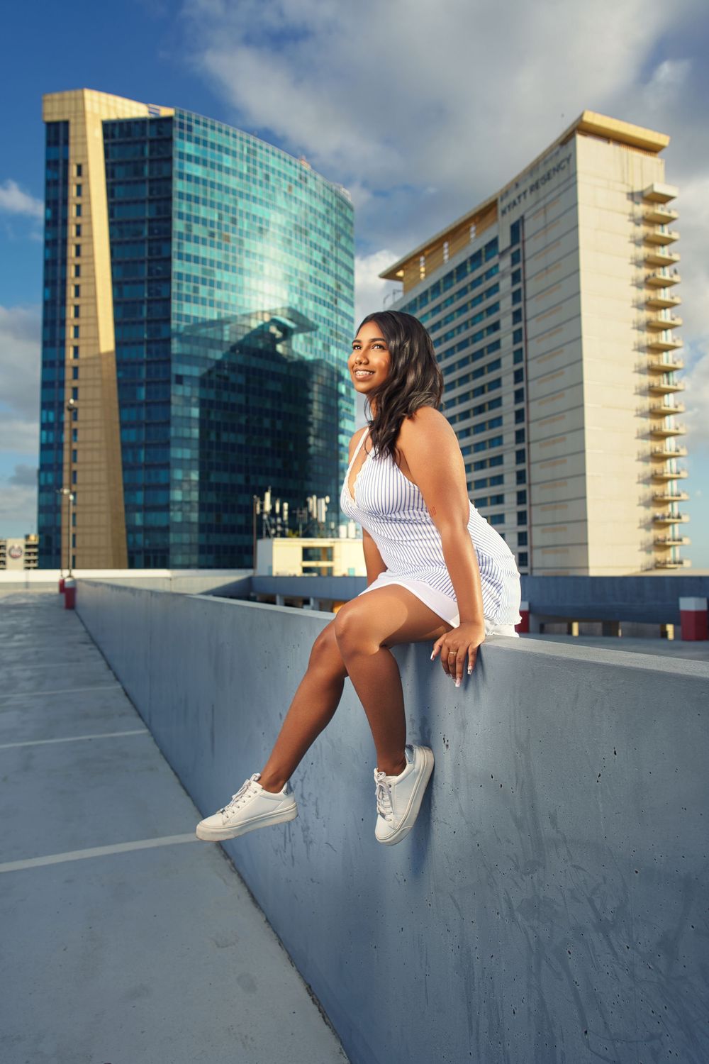 A woman in white athletic wear sits on a curved concrete wall with modern high-rise buildings in the background.