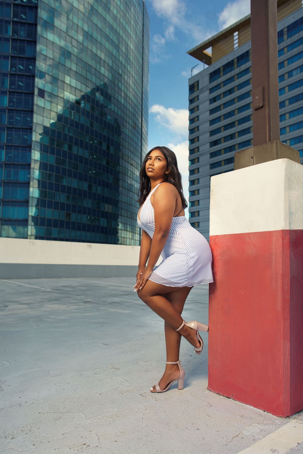 Model in white dress poses against modern city backdrop with glass buildings and red architectural elements.