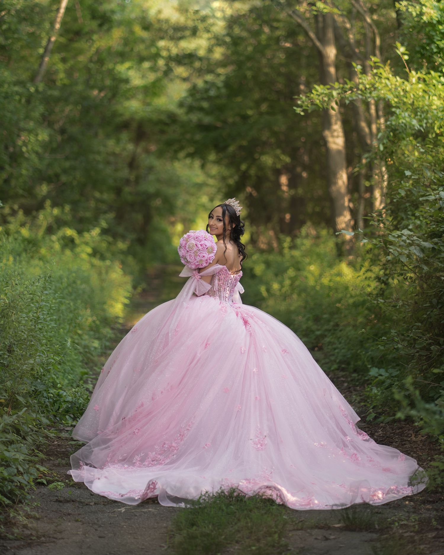 A flowing pink princess ball gown dress photographed in a lush green forest setting creates a dreamy fairytale scene.