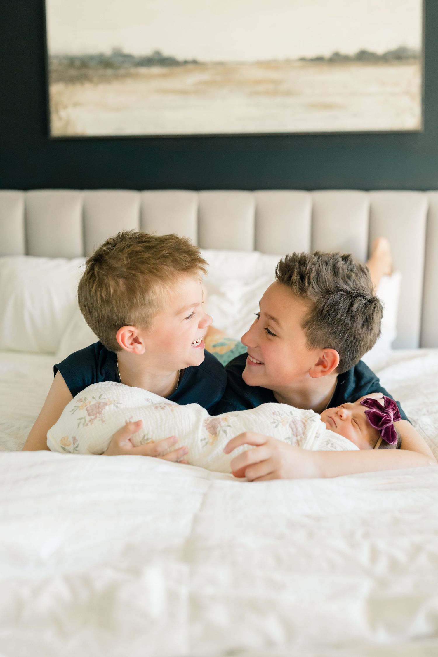 Two young boys lying on white bedding share a playful laugh while facing each other.