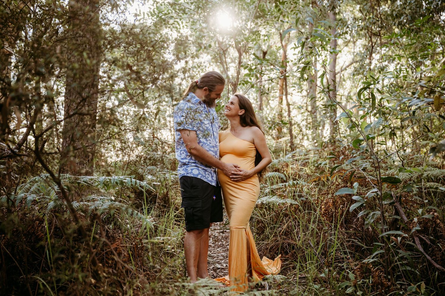 A romantic couple shares tender moments in a sunlit forest setting during an outdoor photo session.