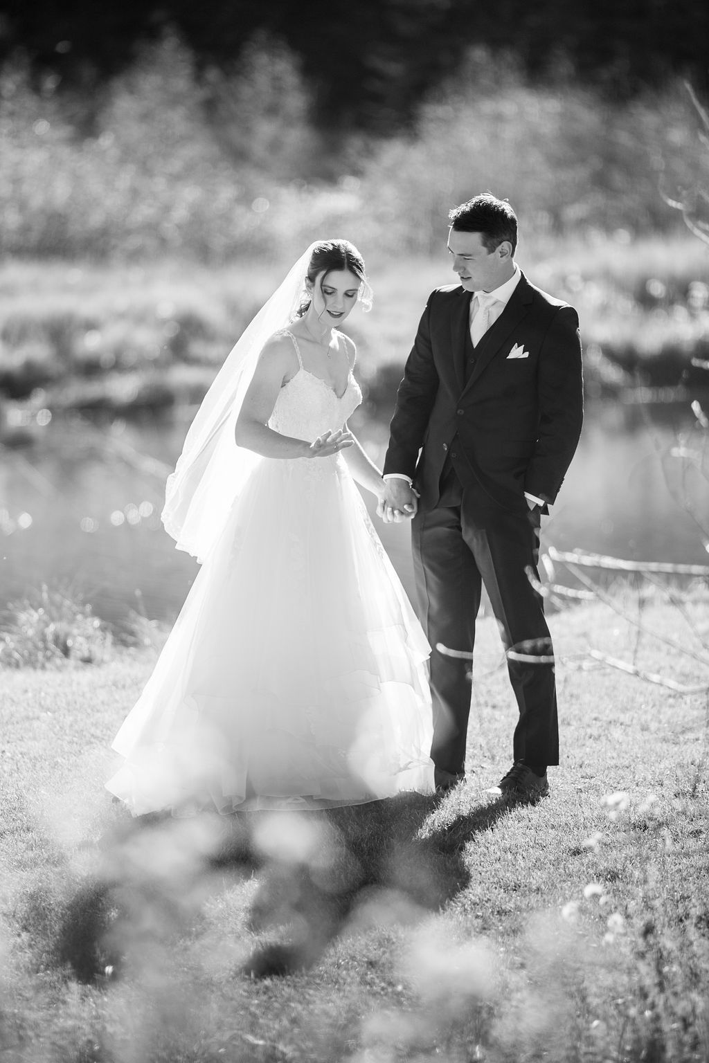 A black and white wedding photo showing a couple outdoors by a lake, with the bride in a flowing white gown and veil.