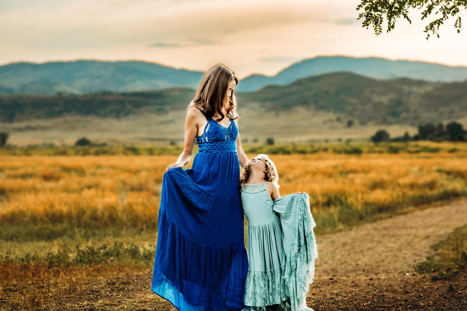Two people in blue dresses walk together along a scenic mountain trail at sunset.