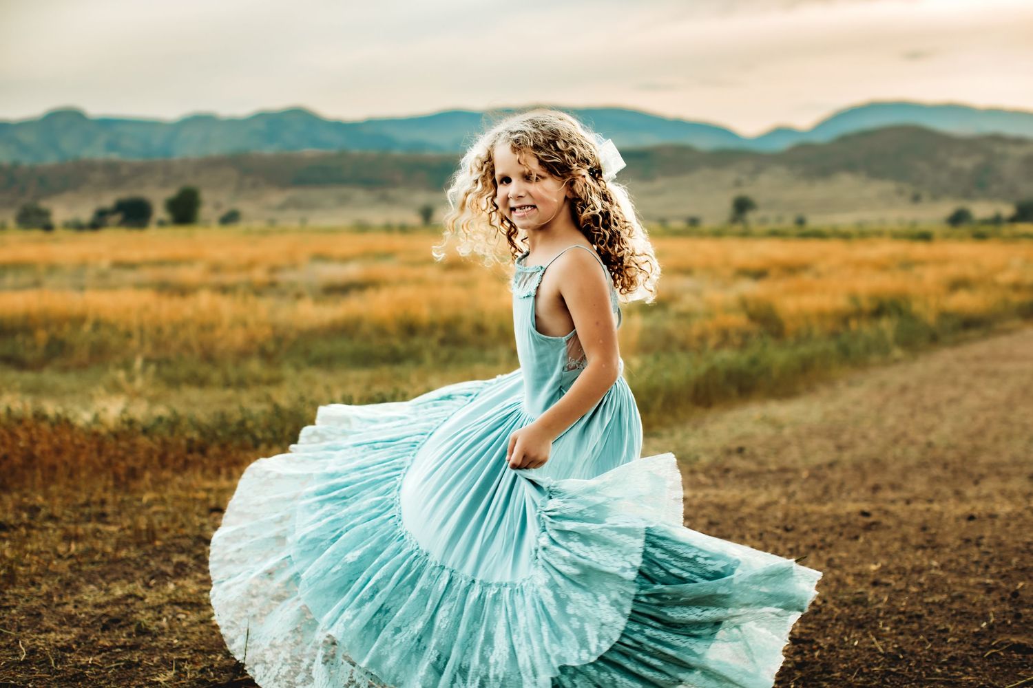 A twirling figure in a flowing mint green dress dances in a golden field with mountains in the background at sunset.