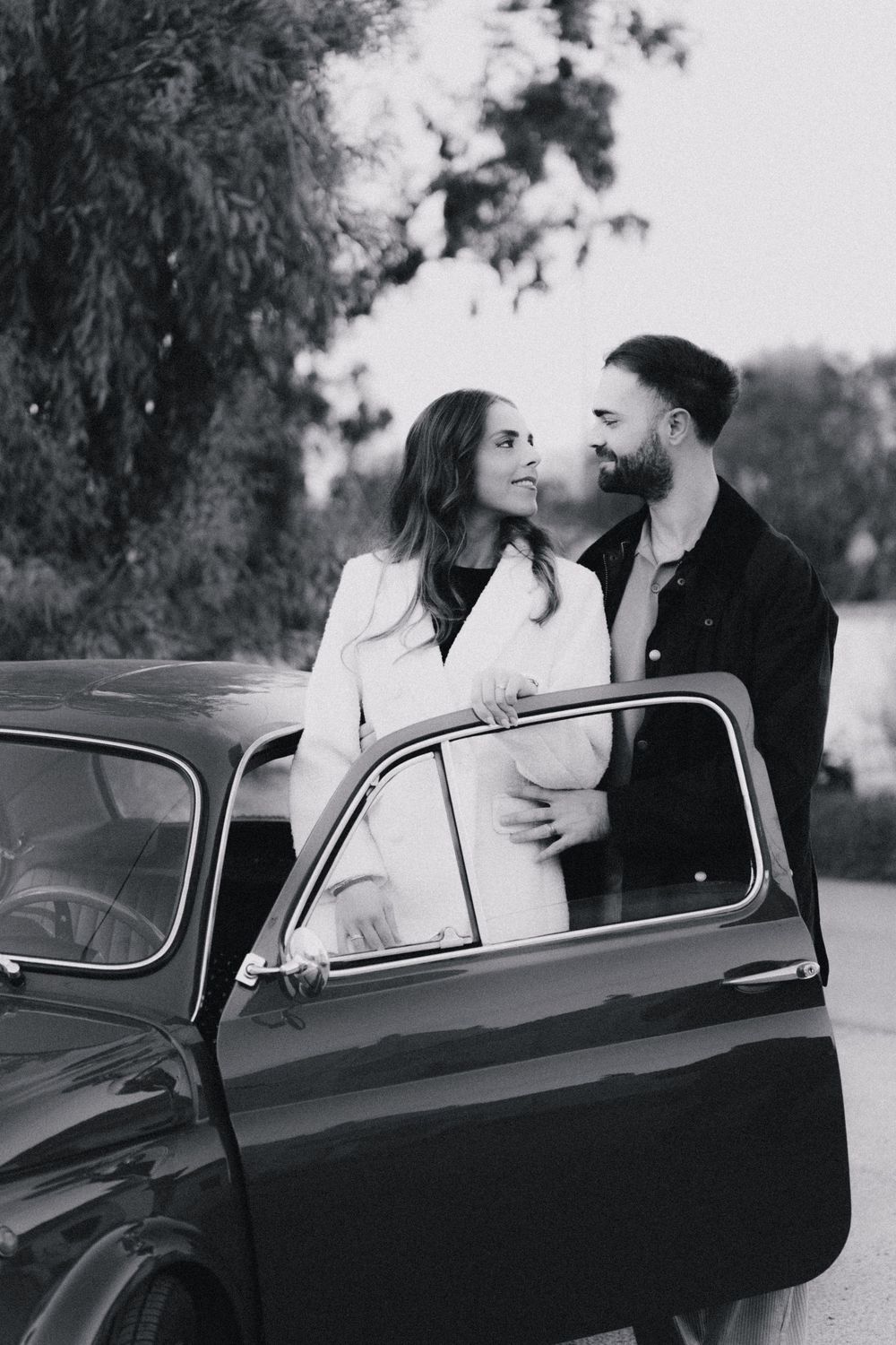 A romantic black and white photo of a couple sharing a tender moment by a vintage car with its door open.