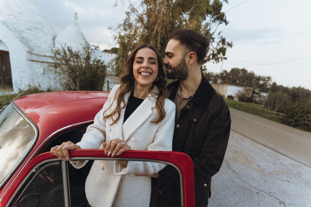 A couple shares intimate moments while posing with a vintage red car during a romantic outdoor photoshoot.