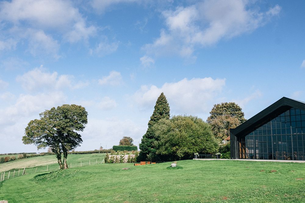 Modern barn venue with glass walls sits on a lush green hillside under blue skies with scattered white clouds.