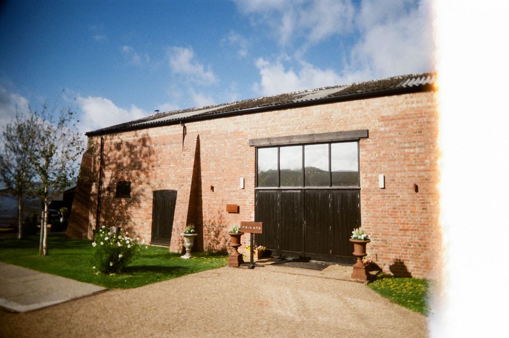 Panoramic view of a rustic brick barn venue with large windows and landscaped grounds under blue skies.