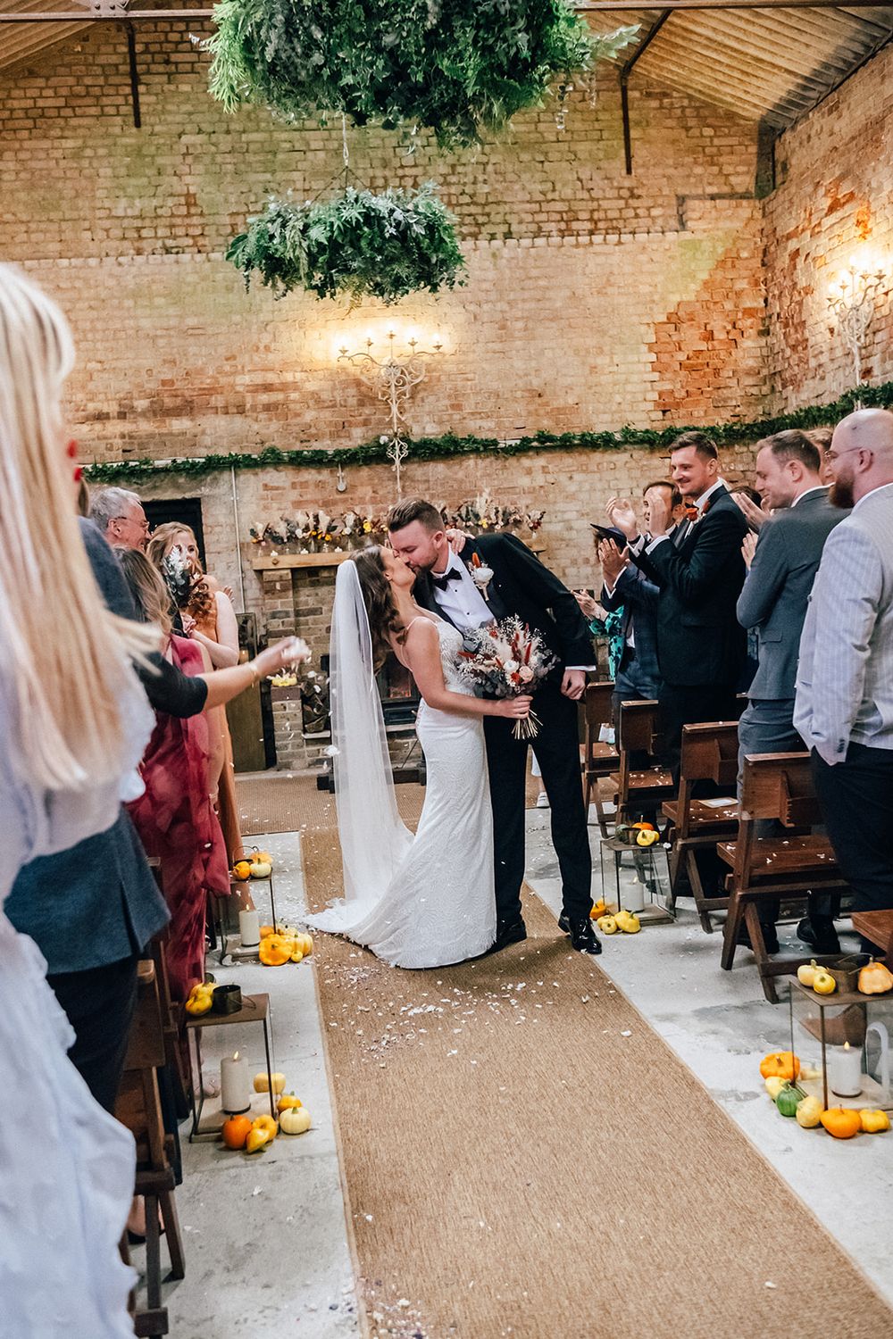 Wedding guests throw confetti at a couple during a reception celebration in a rustic brick-walled venue with pumpkin decor.