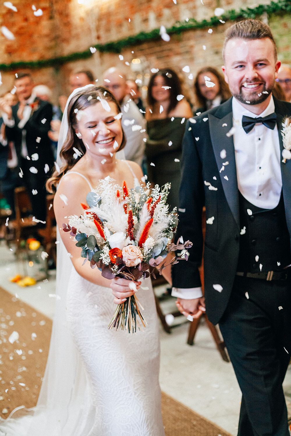 A joyful wedding celebration with confetti falling as the couple walks together, the bride holding a red and white bouquet.