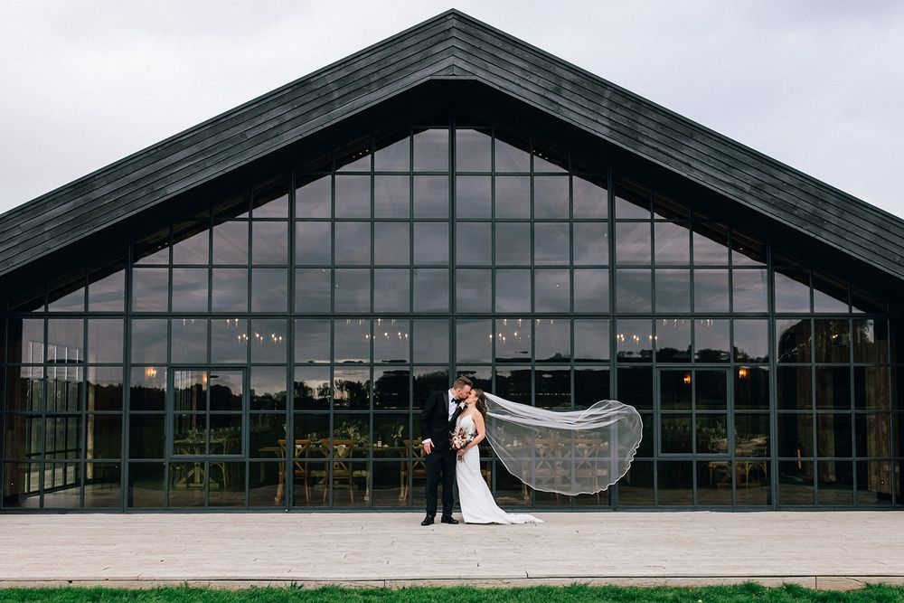 The Main Barn - Wedding couple poses in front of modern glass and steel building for dramatic panoramic shot at sunset.