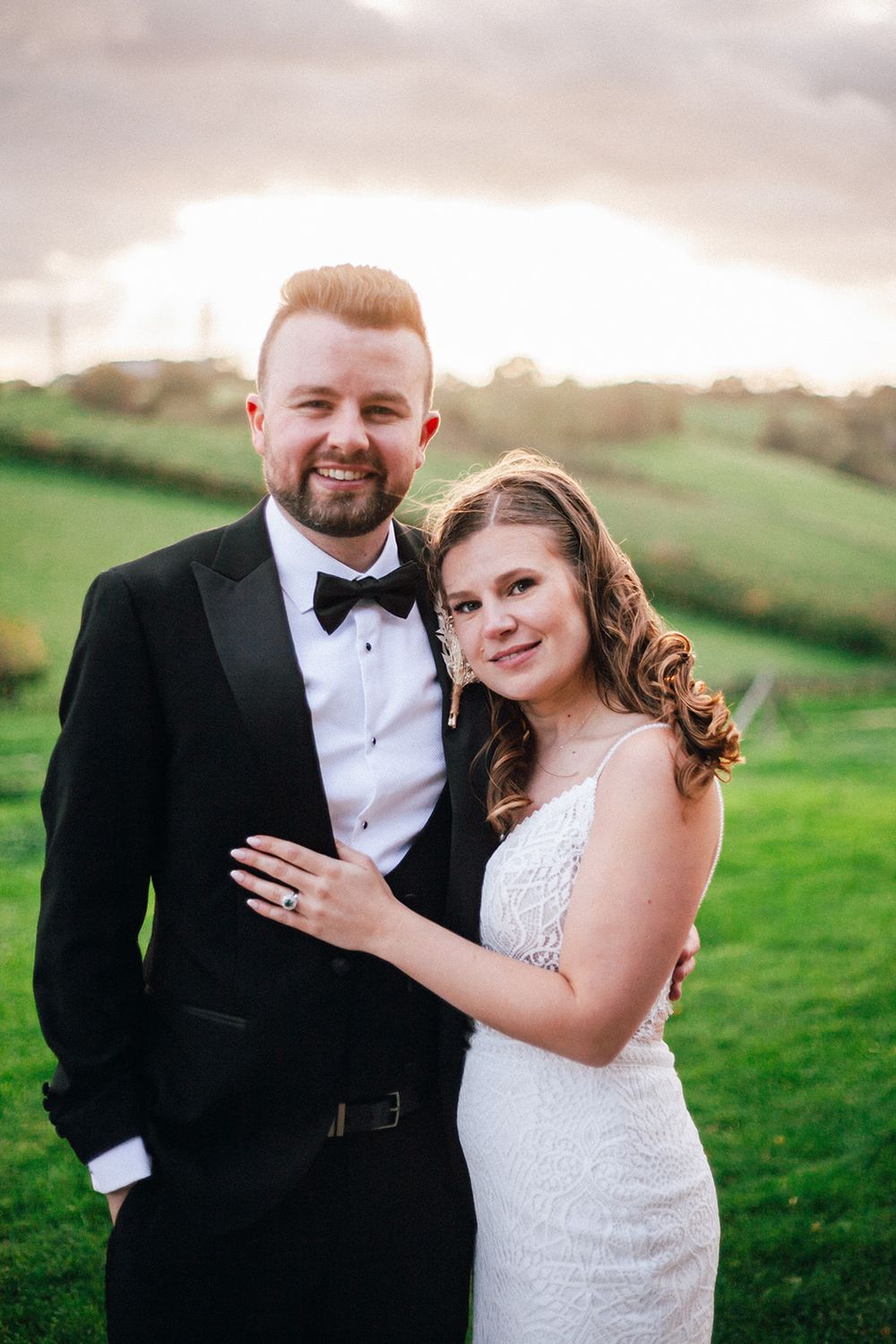 Surrey Wedding Venue - Botley Hill Barn. A newlywed couple poses outdoors with green rolling hills and a sunset sky behind them.