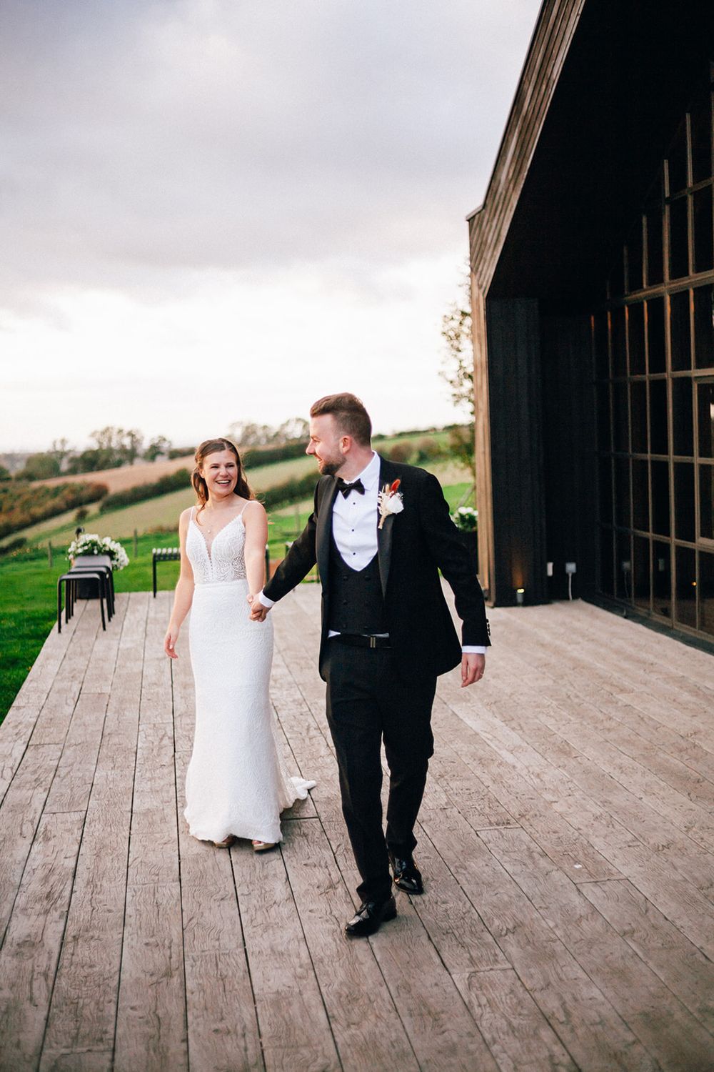 A couple walking together on a rustic wooden deck with rolling hills in the background on their wedding day.