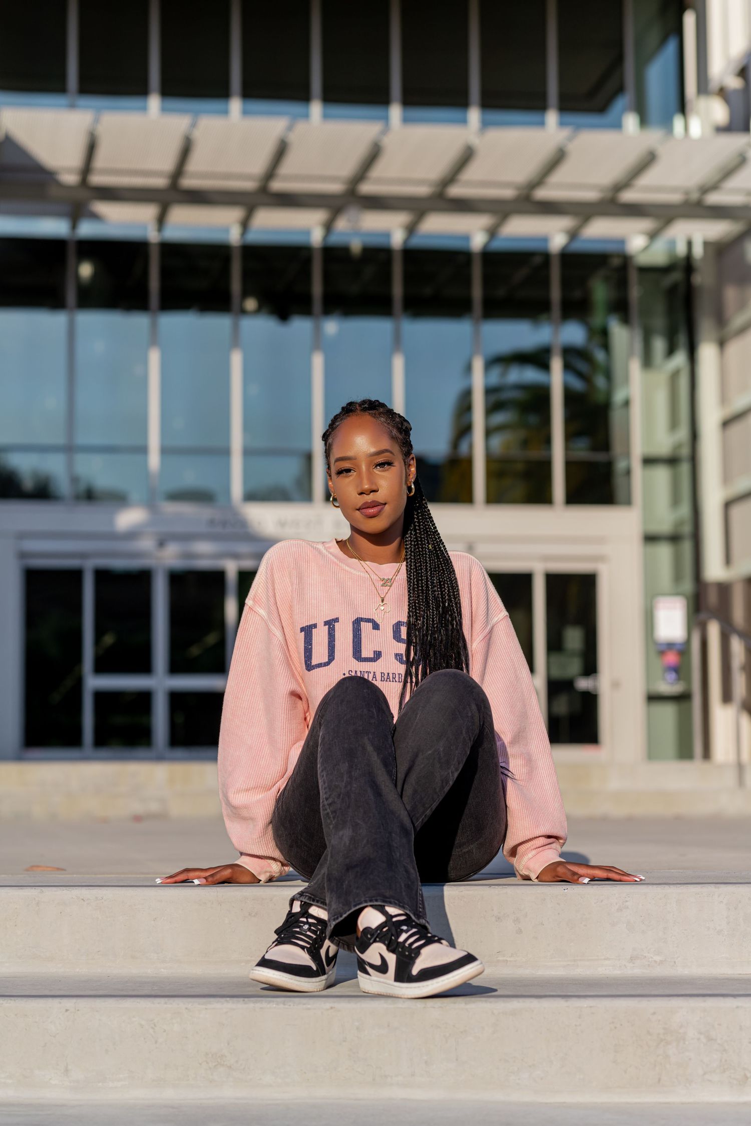 A person in a pink UCLA sweatshirt and jeans sits casually on concrete steps outside a modern building with glass walls.