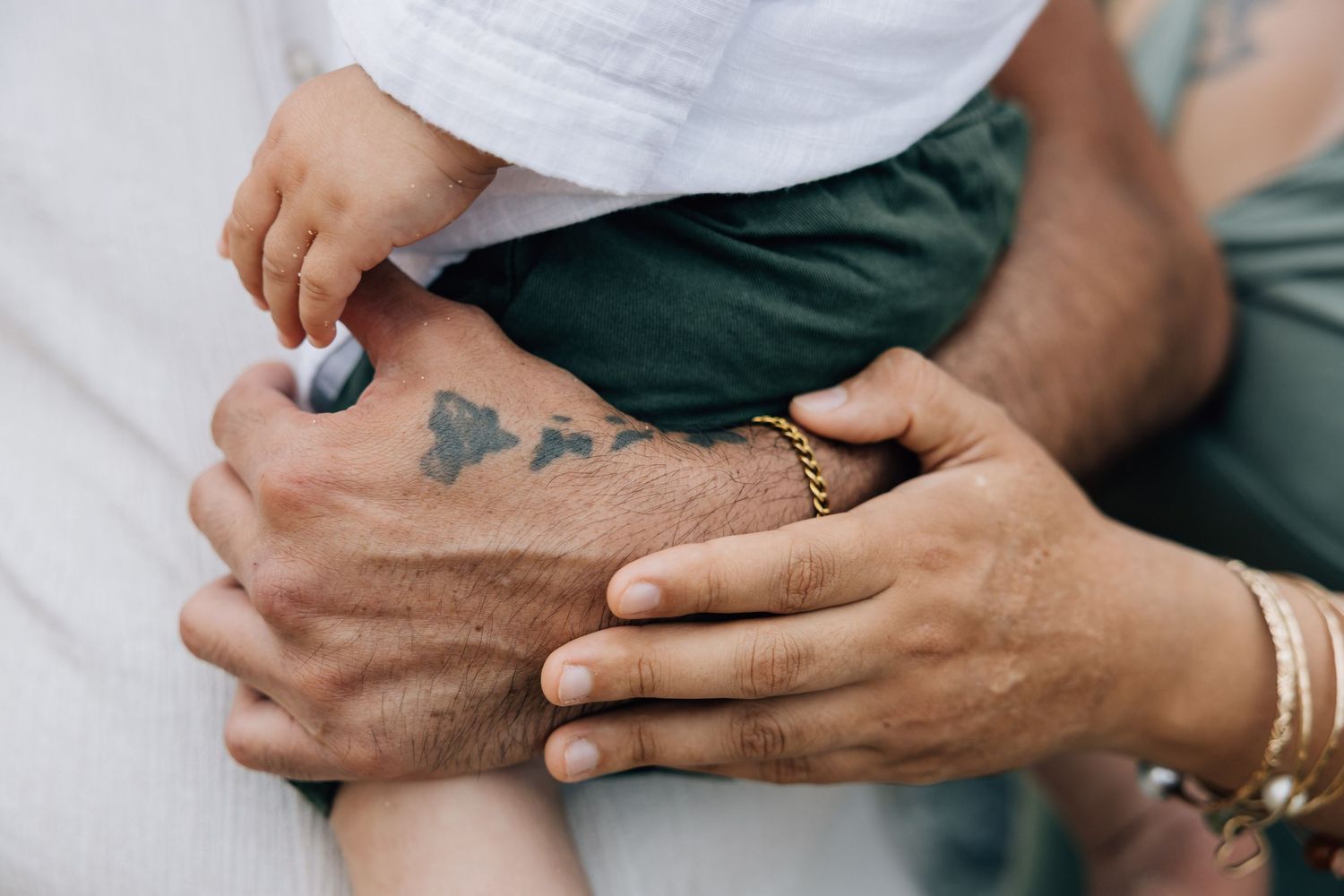 Close-up of father holding baby with tattoo and gold bracelet visible.
