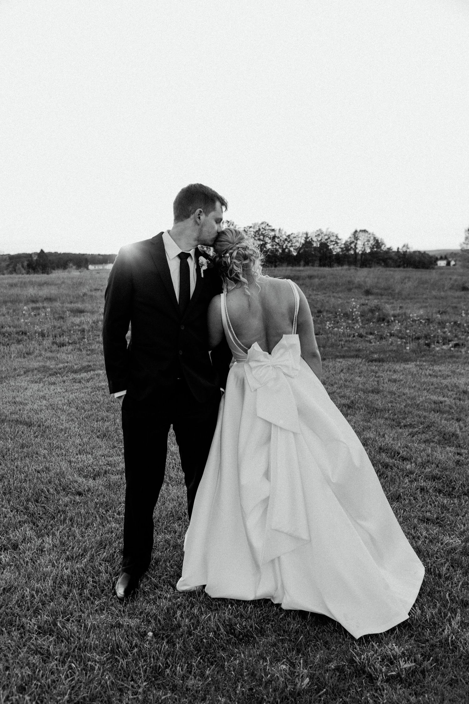 A black and white wedding portrait shows a couple in formal attire standing in an open field.