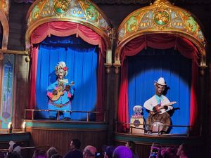 Animatronic musicians perform with guitars in ornate theater booths with blue curtains and decorative gold arches.