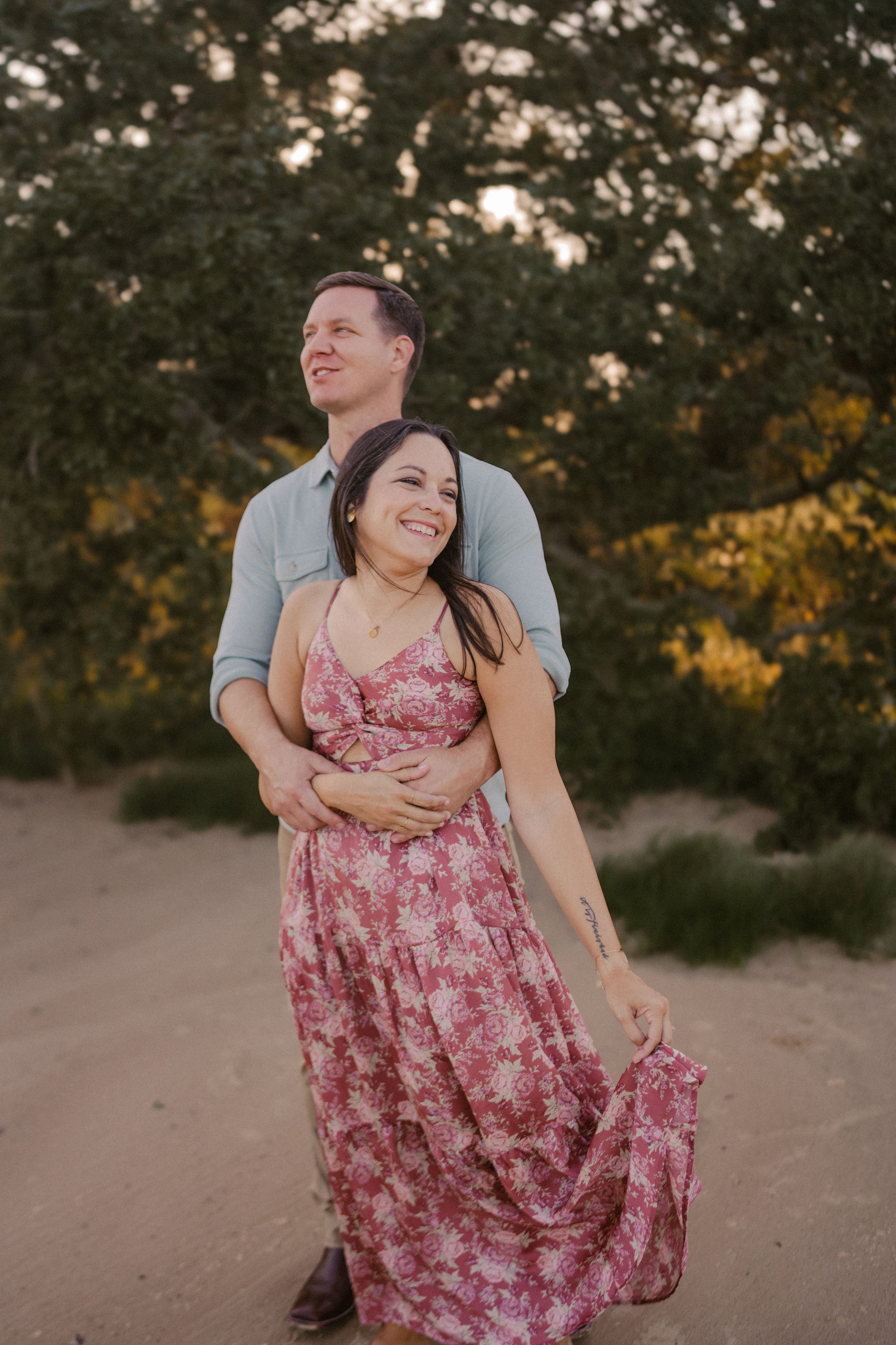 Couple embraces on sandy trail during sunset as woman in floral pink dress smiles joyfully.