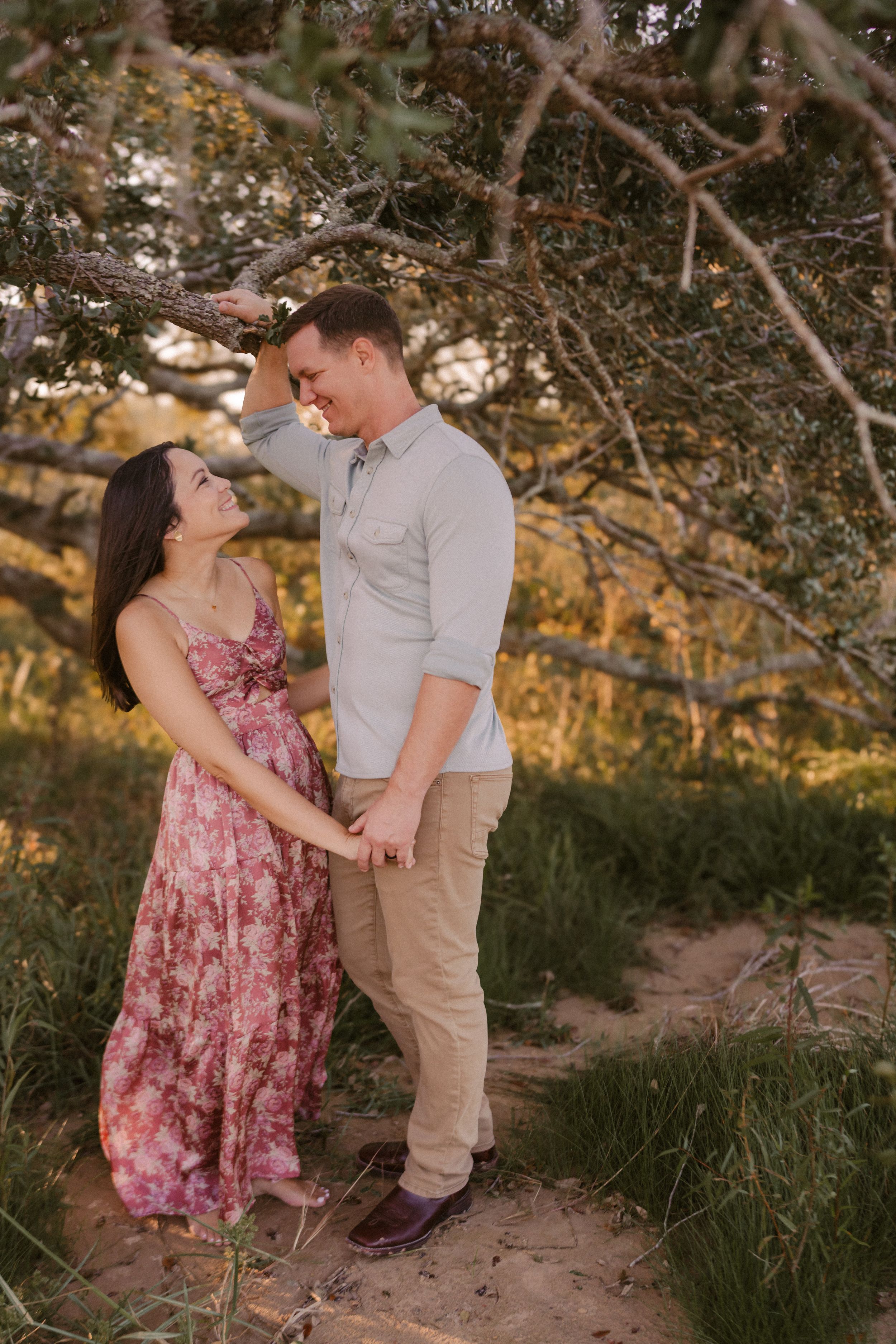 A couple shares an intimate moment on a sandy path under tree branches at golden hour.