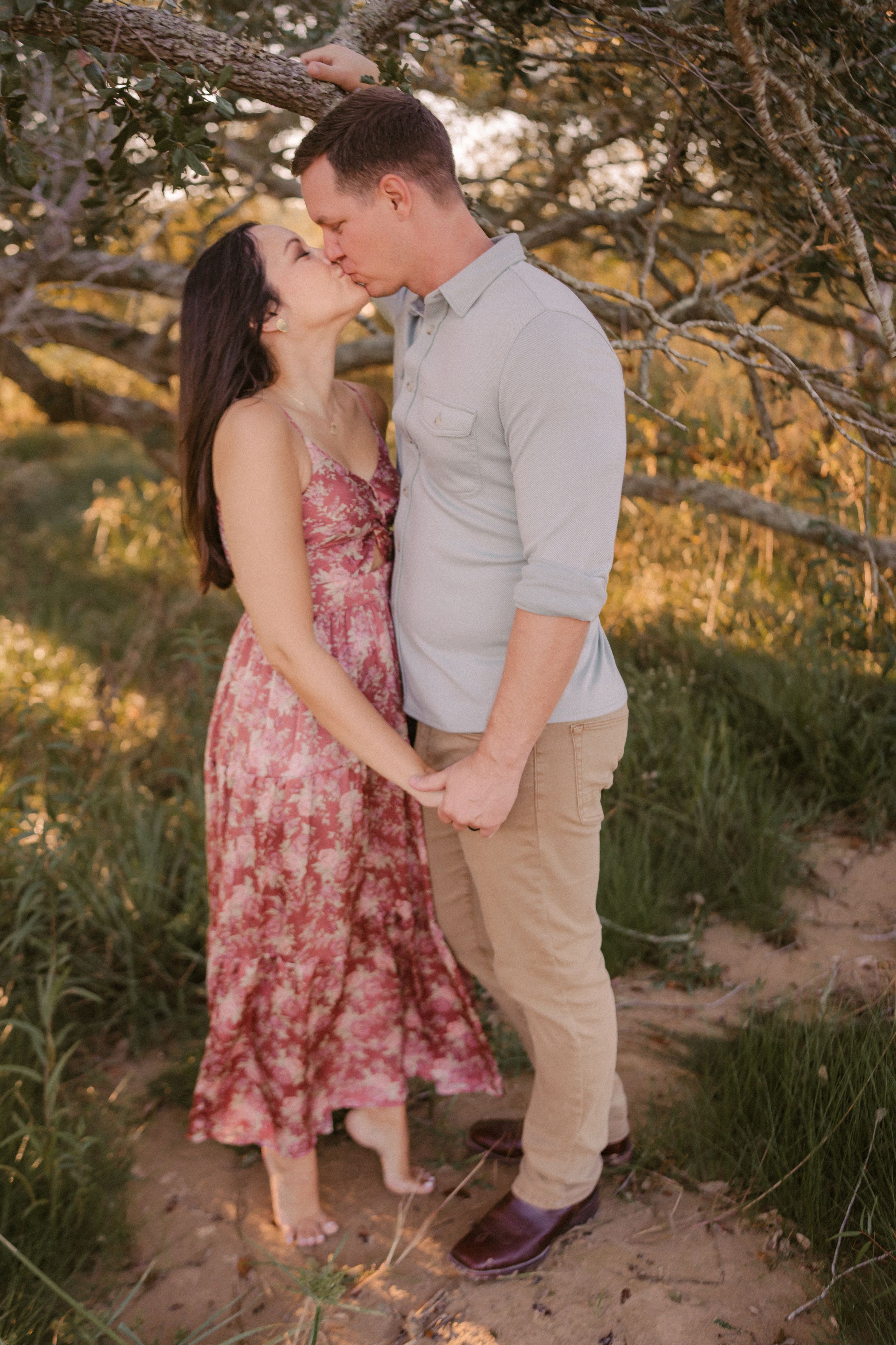A couple shares a romantic moment outdoors in golden light, with her in a pink floral dress and him in a light button-down shirt.