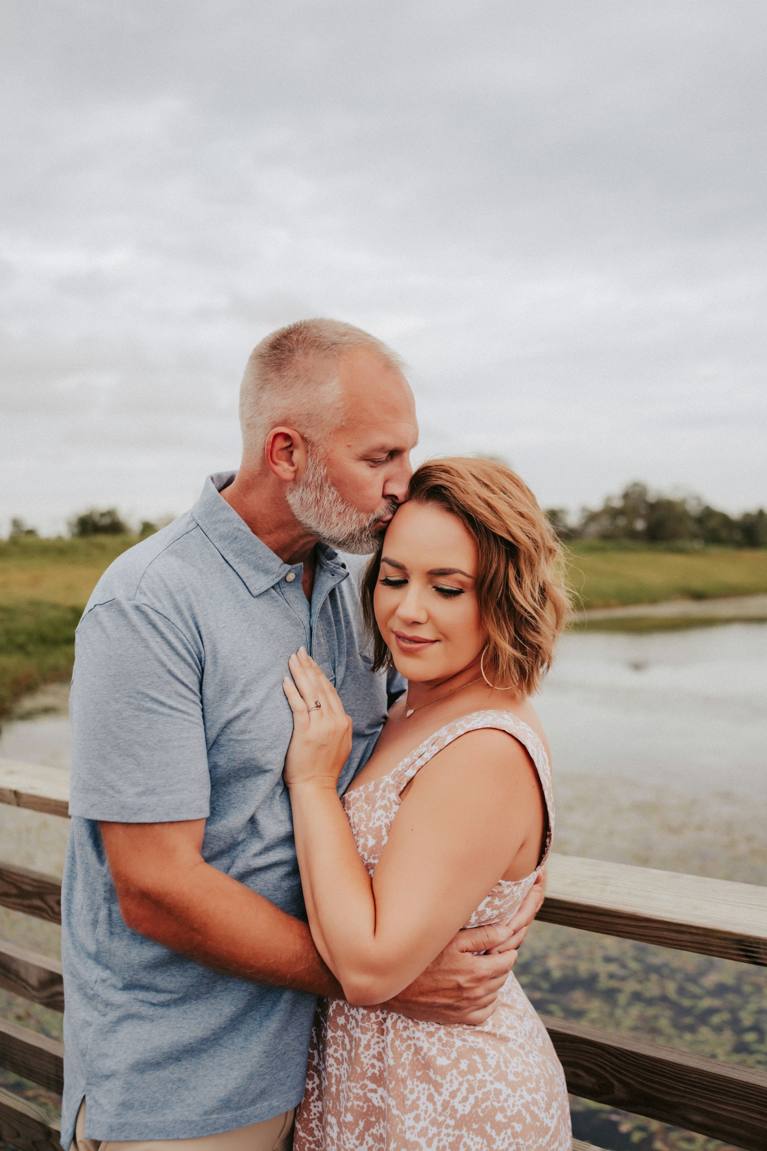 Couple embraces tenderly on a wooden dock overlooking water during romantic outdoor photoshoot at dusk.