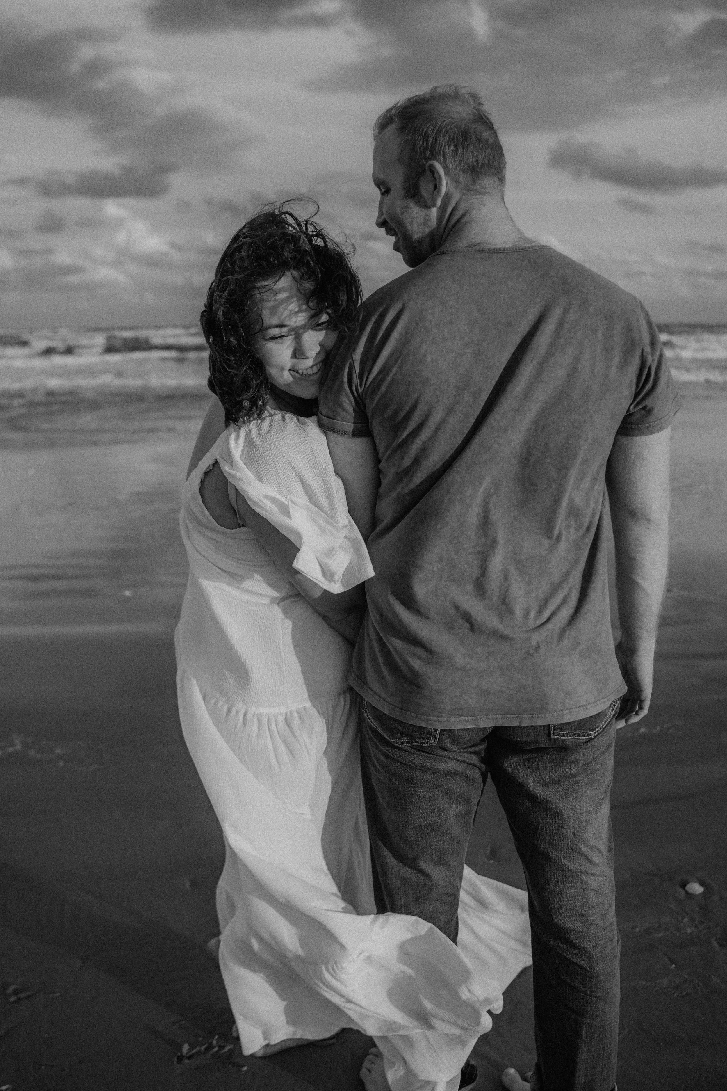 A couple embraces on a moody black and white beach, with waves crashing and cloudy skies in the background.