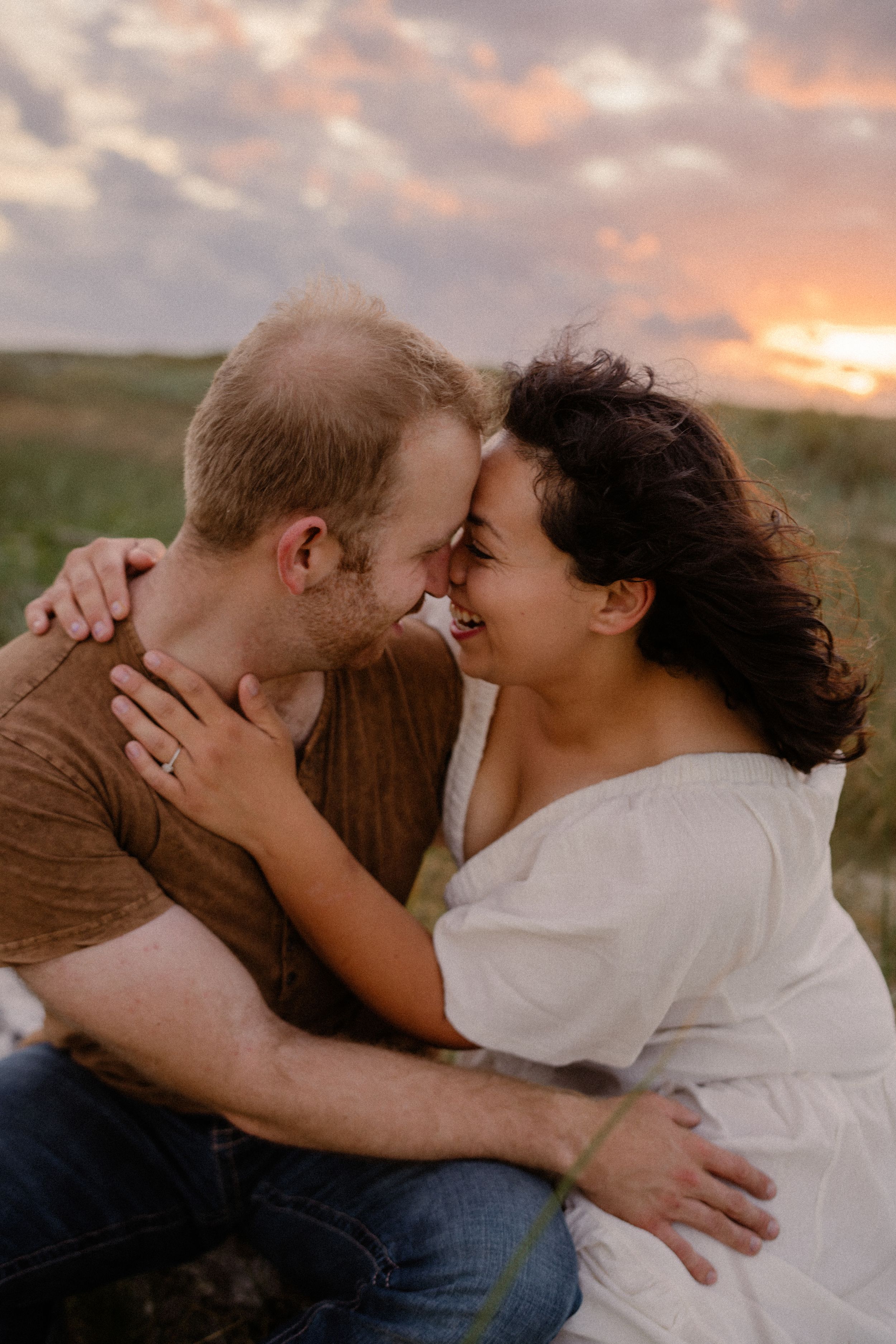 A couple embracing and laughing together during sunset in a grassy field, with warm orange clouds in the sky.