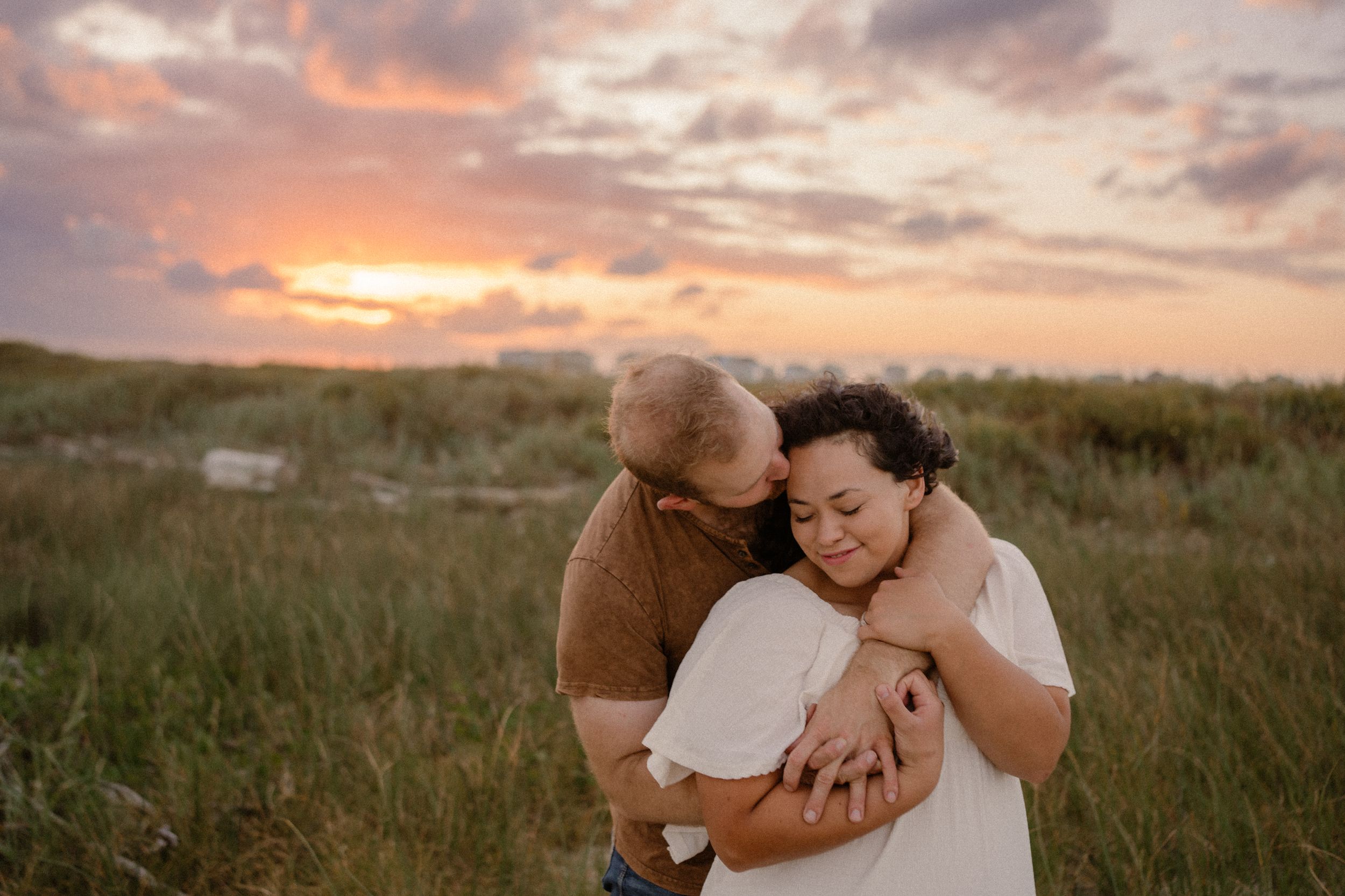 Couple embraces tenderly in a grassy field during golden sunset with dramatic clouds in the sky.