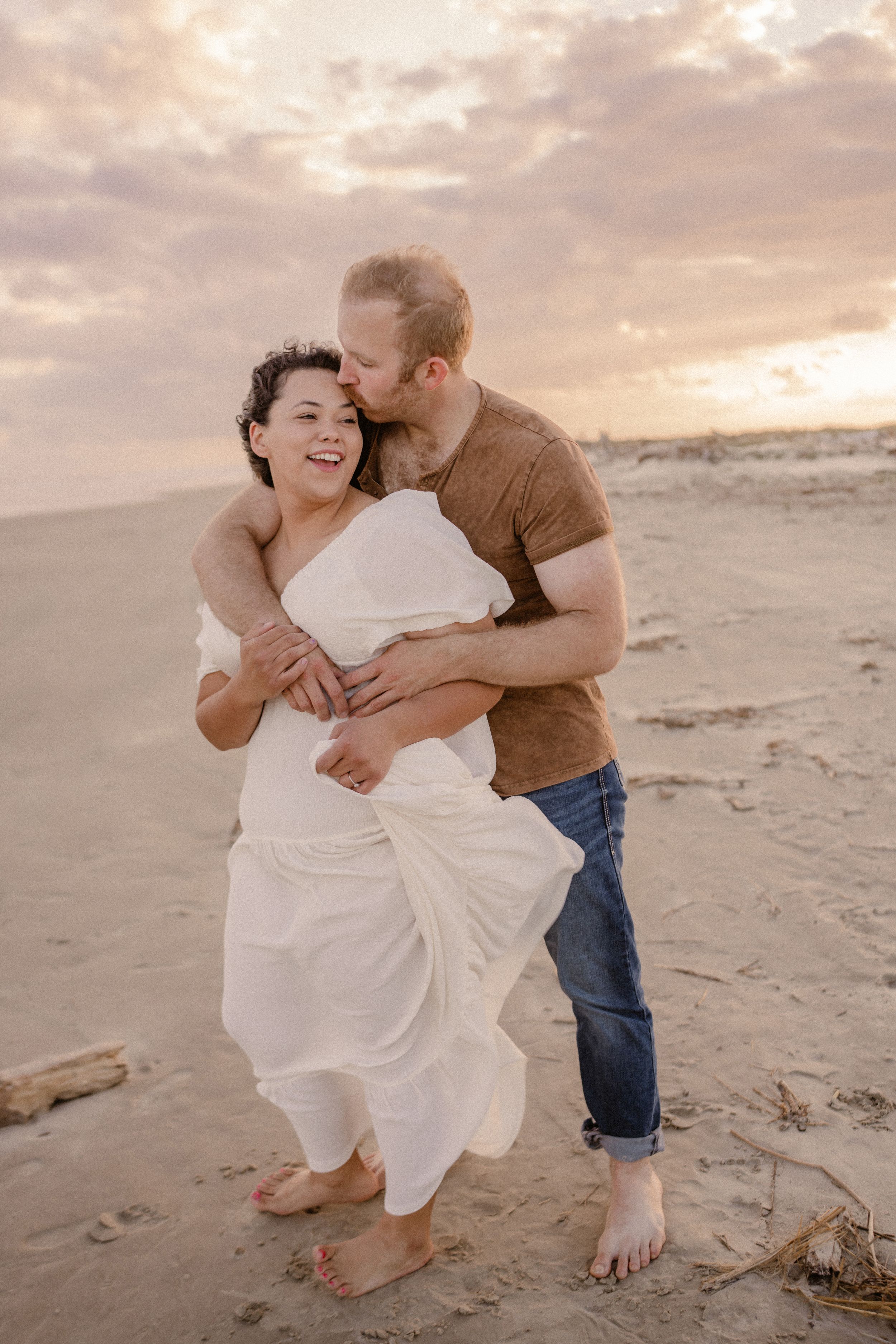 A couple embraces and laughs together on a sandy beach at sunset with soft warm lighting.
