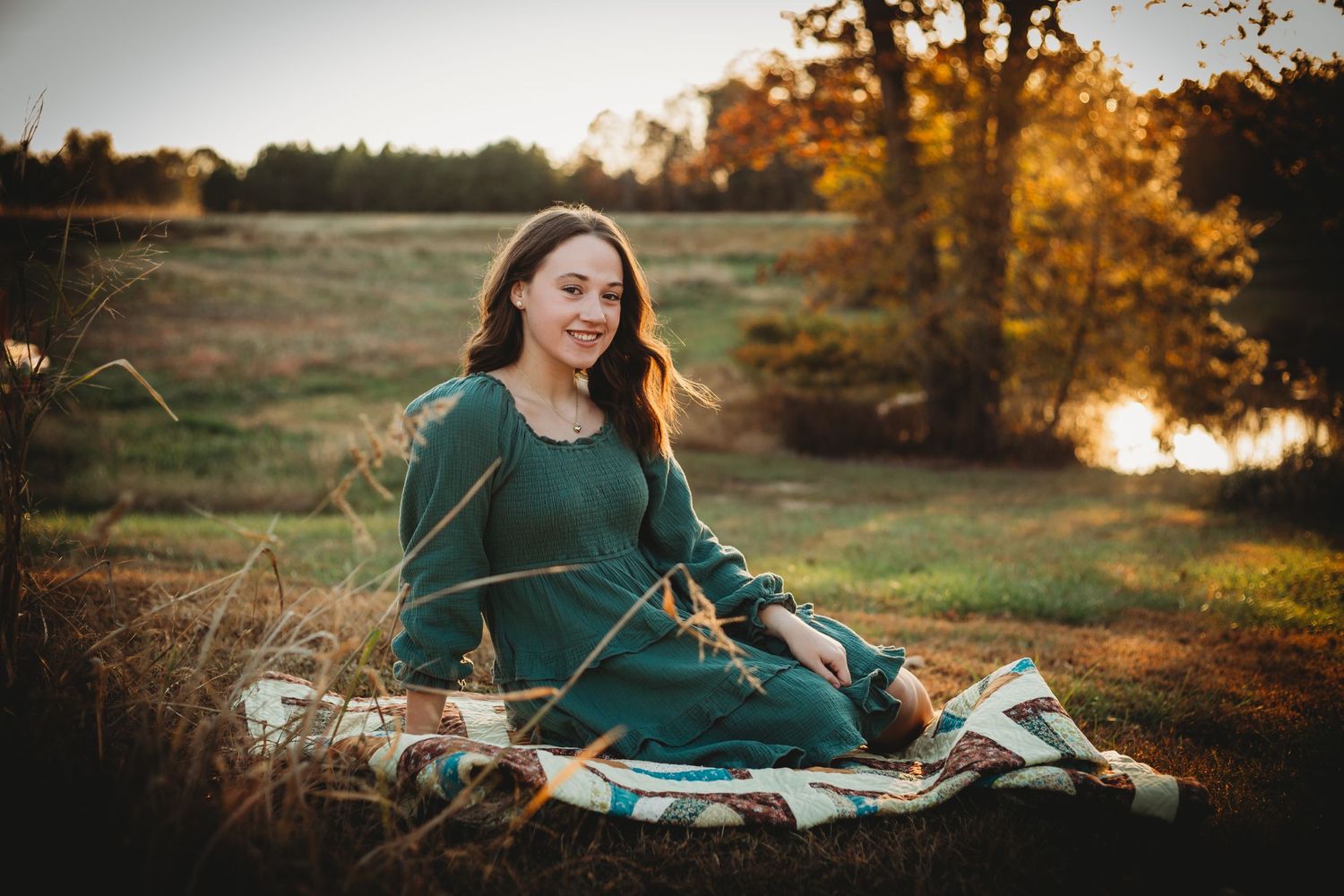 A person in a green dress sits on a blanket by a pond during golden hour sunset with autumn trees in the background.