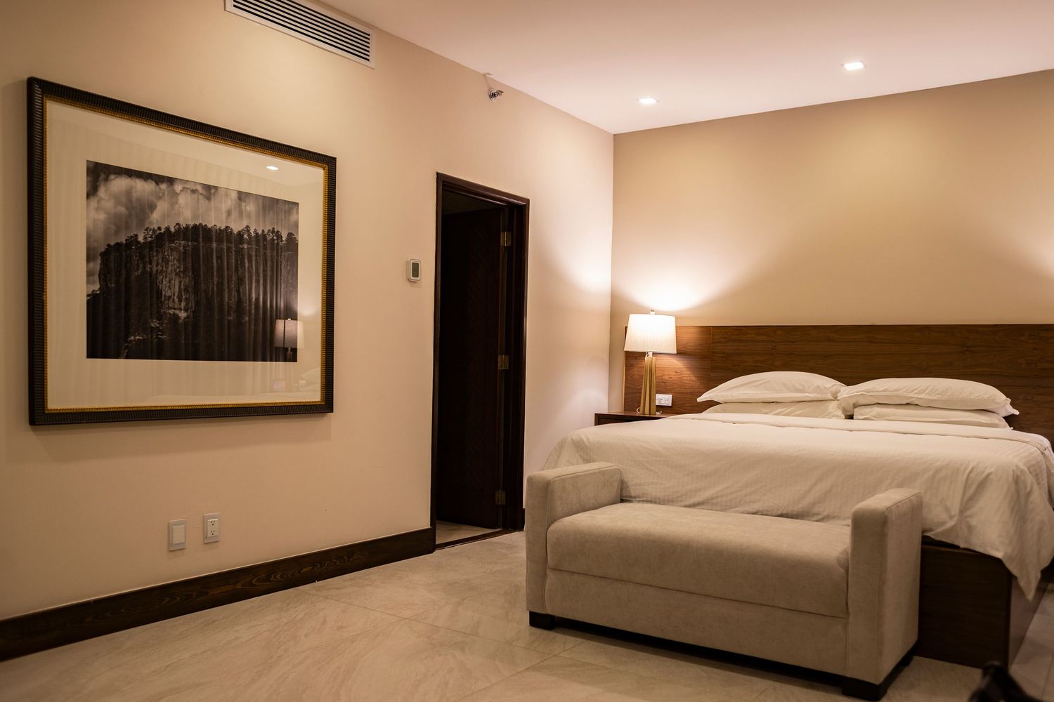 Elegant hotel bedroom with a large bed, bedside lamp, framed black-and-white artwork, and a bench at the foot of the bed.