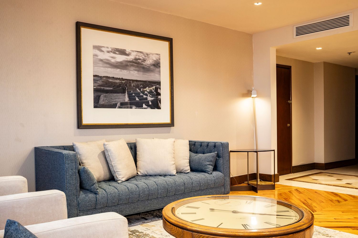Elegant hotel suite living area with blue sofa, decorative pillows, large clock coffee table, and black-and-white cityscape artwork.