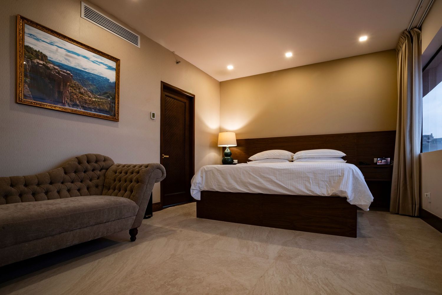 Elegant hotel room with king bed, tufted sofa, warm lighting, landscape painting, and neutral tones.