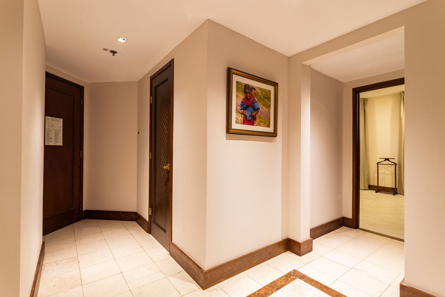 Bright hotel hallway with beige walls, tile floors, dark wood doors, and colorful framed artwork near the entrance.