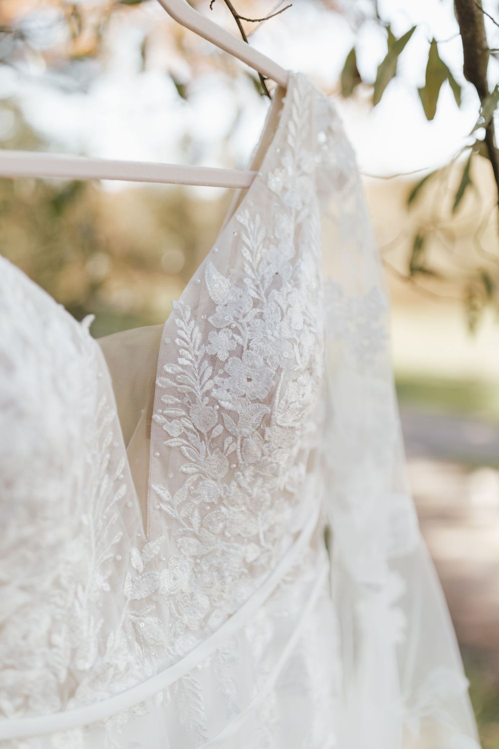 Close-up detail of a white lace wedding dress with intricate floral patterns hanging outdoors.