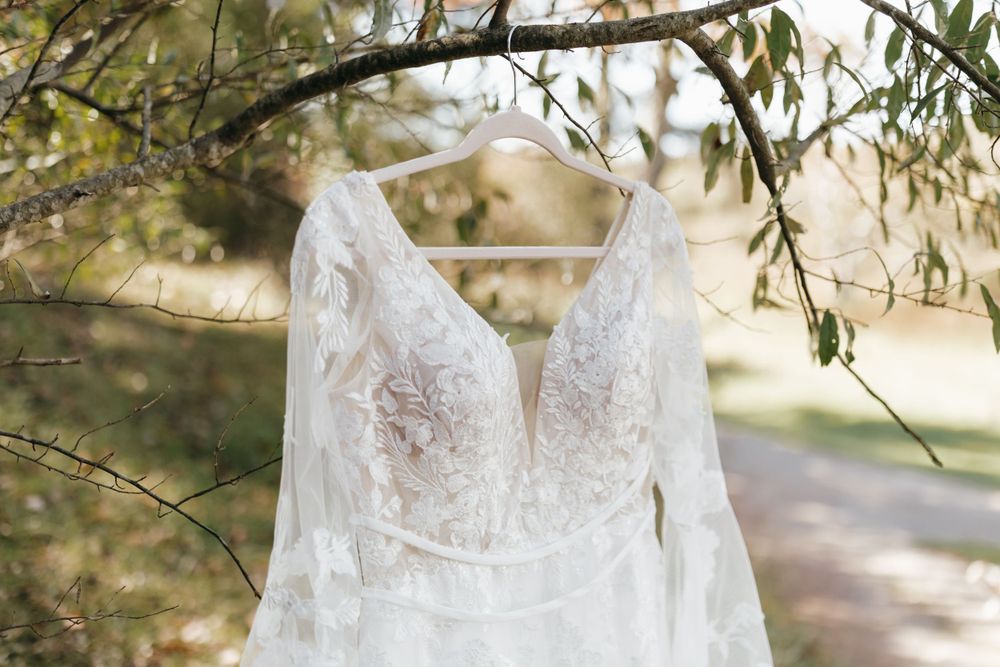 Elegant white lace wedding dress with plunging neckline hangs from a tree branch in natural lighting.
