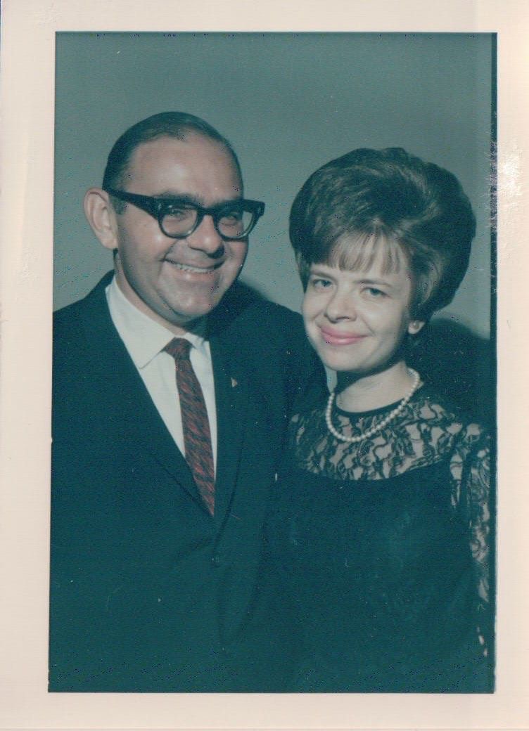 A vintage 1960s portrait photograph shows a couple posing together, with the man wearing glasses and a tie.