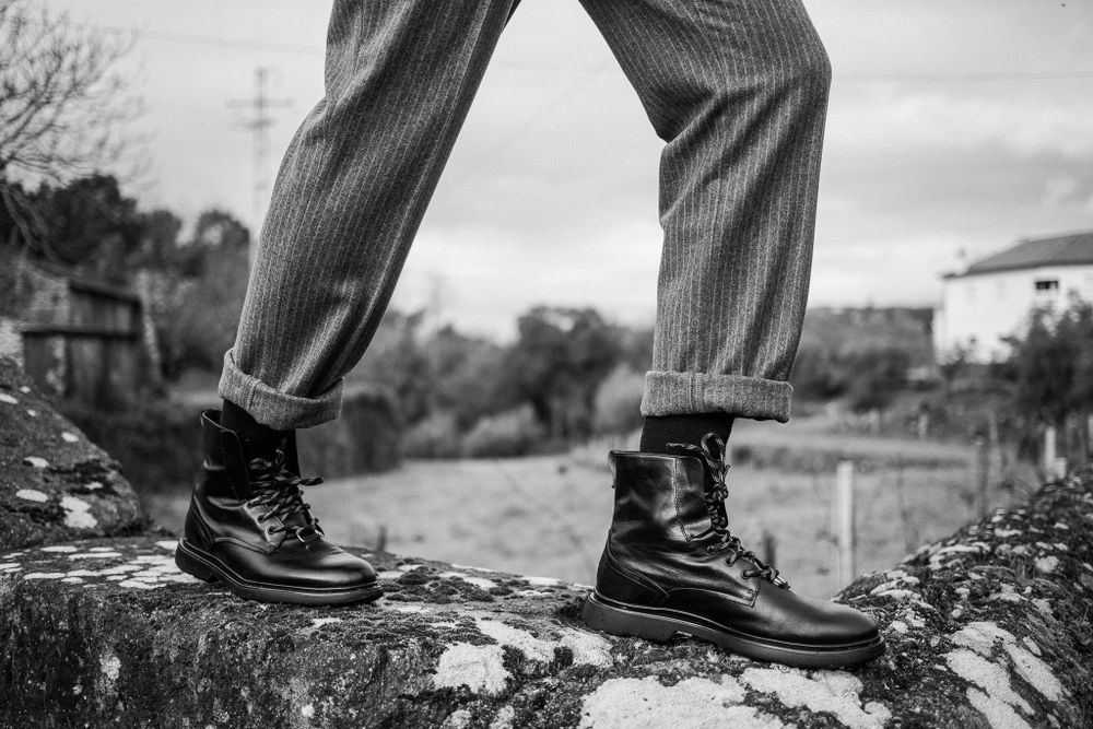 Black and white photo of rolled-up pinstripe pants and black leather boots standing on rugged stone surface outdoors.