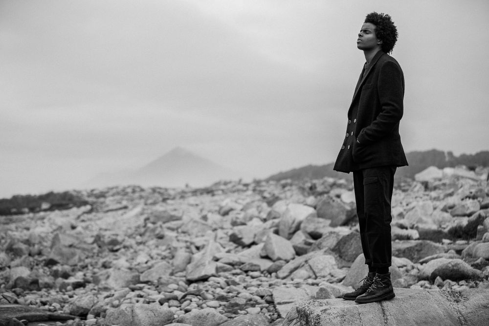 Black and white photo of someone standing contemplatively on rocky terrain against a misty mountainous backdrop.