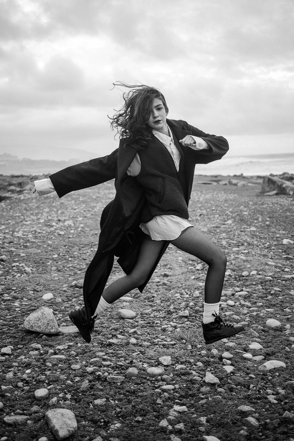 Black and white photo of person in flowing coat dancing dramatically on rocky terrain under cloudy sky.