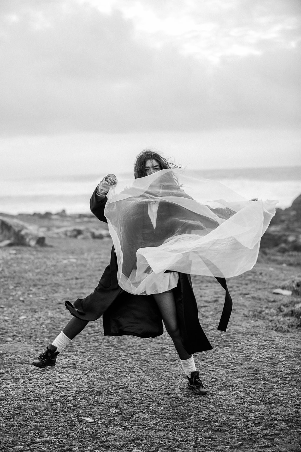 Black and white artistic dance photo capturing movement with flowing white fabric against cloudy coastal landscape.