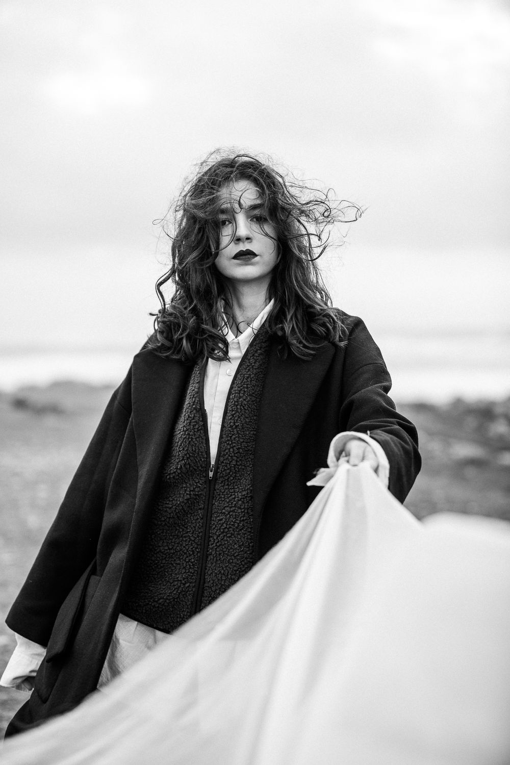 A dramatic black and white portrait in moody lighting shows windswept hair against cloudy skies while holding flowing fabric.