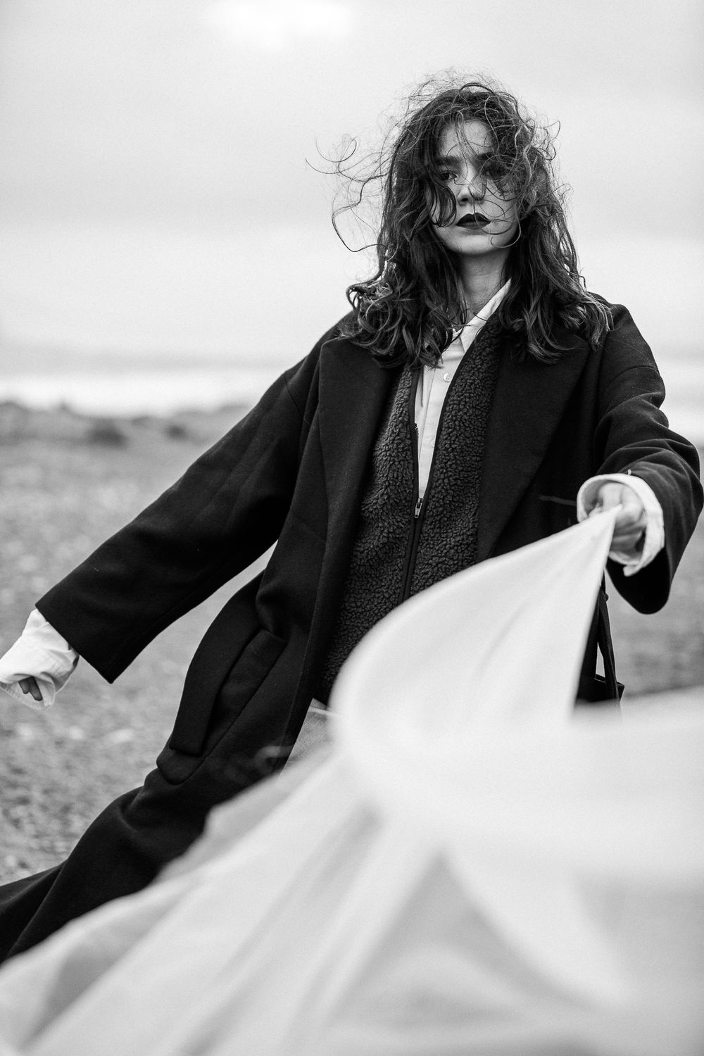 Dramatic black and white fashion photo showing figure in long dark coat with windswept hair holding flowing white fabric.