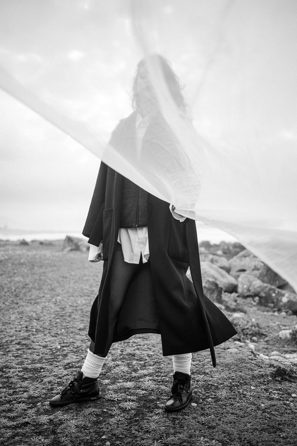 A figure in a flowing black coat and white socks stands on rocky terrain, with a translucent veil creating a dreamy effect.