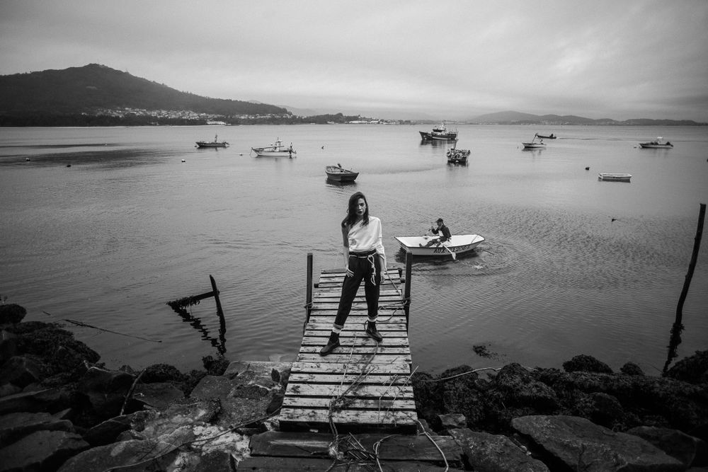 A dramatic black and white photo shows a figure standing on a wooden pier overlooking boats in a misty harbor.