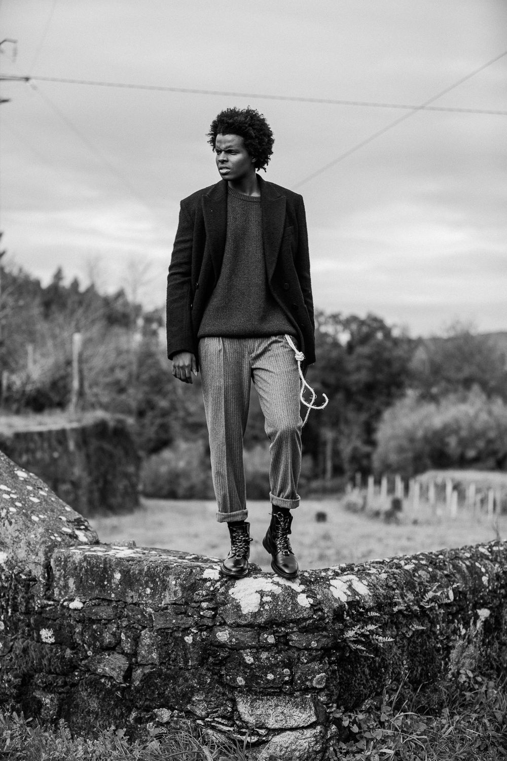 Black and white portrait of figure in dark blazer and pants standing on stone wall with rural landscape background.