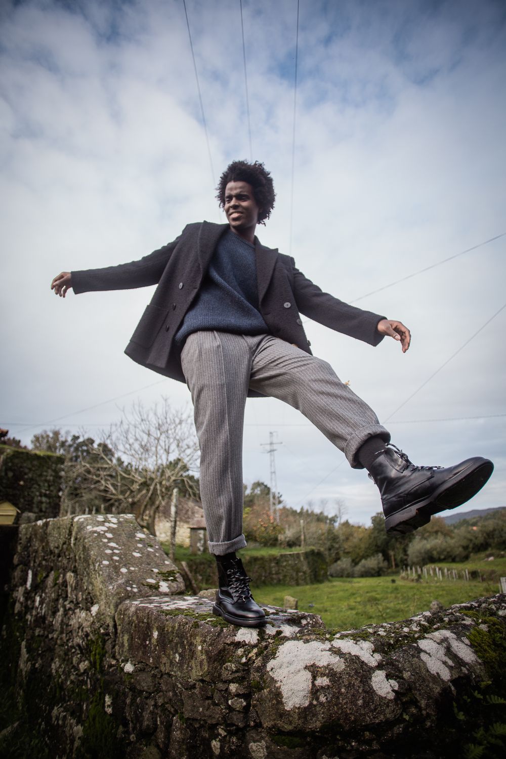 A person balancing on a stone wall wearing a dark blazer and grey pants against a cloudy sky with power lines.